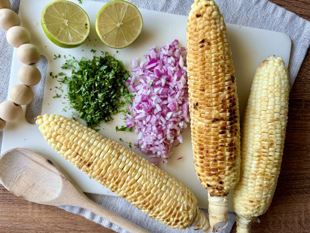 roasted corn on the cob, chopped red onion and chopped cilantro sitting on a table with lime 