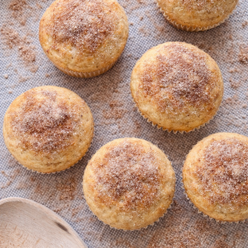 overhead shot of churro kodiak protein muffins with cinnamon sugar sprinkled around them on a mat next to a wooden spoon
