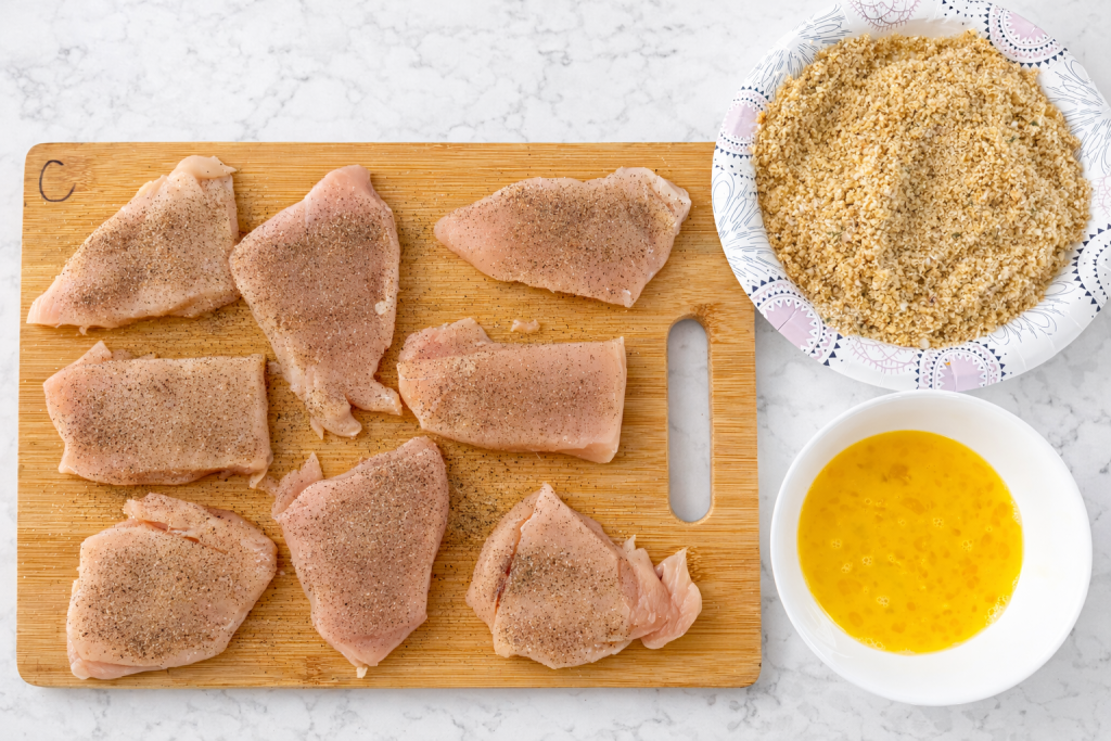 chicken breast pieces on cutting board next to a bowl with egg wash and another bowl with breadcrumbs