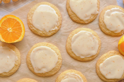 orange creamsicle cookies topped with glossy vanilla-orange icing. The cookies have a soft, homemade texture with lightly golden edges and subtle cracks. Surrounding the plate are fresh orange halves
