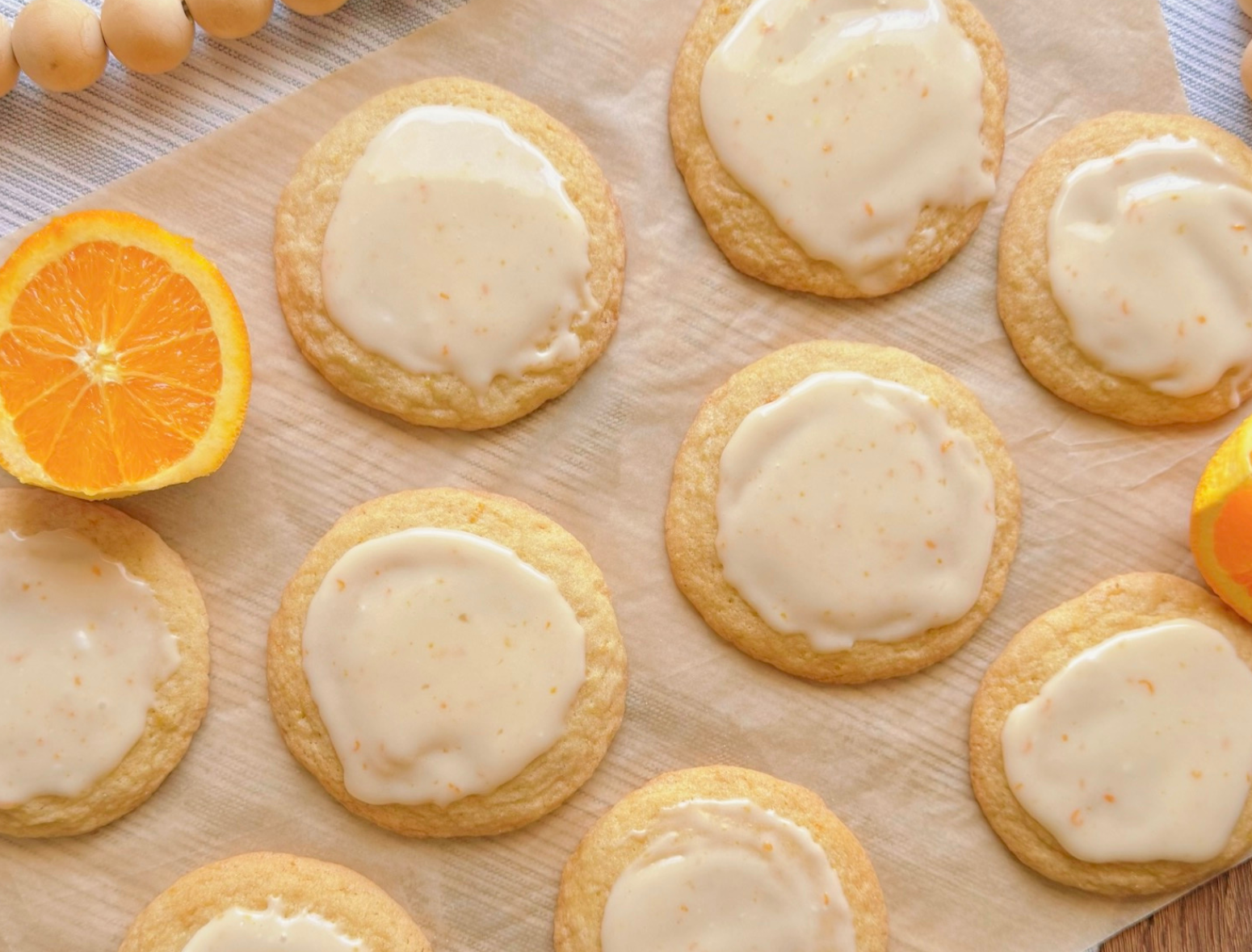 orange creamsicle cookies topped with glossy vanilla-orange icing. The cookies have a soft, homemade texture with lightly golden edges and subtle cracks. Surrounding the plate are fresh orange halves